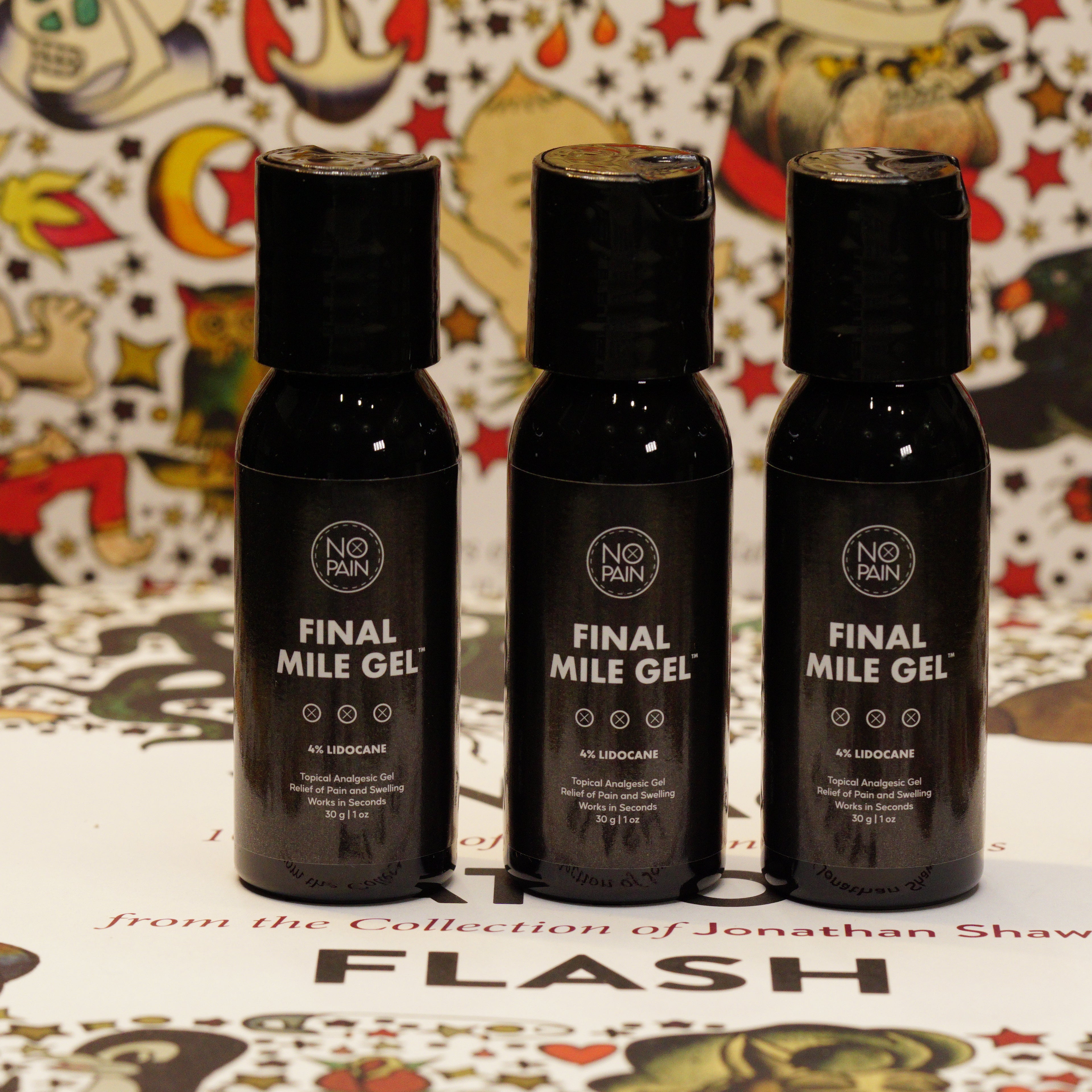 Three sleek black bottles labeled "Final Mile Gelâ„¢" from No Pain Tattoo LLC are standing on a decorative surface. The background features colorful, intricate cartoon drawings, with text below the bottles reading "from the Collection of Jonathan Shaw - FLASH." Infused with a lidocaine formula, this gel ensures no pain during your final mile.