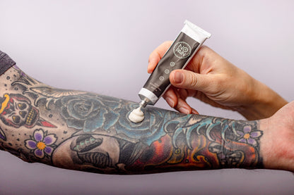 A person applies a generous amount of No Pain Tattoo Numbing Cream from My Store to a heavily tattooed forearm. The tattoo features colorful flowers, roses, and various intricate designs. The background is a plain, light color, ensuring tattoo comfort throughout the process.