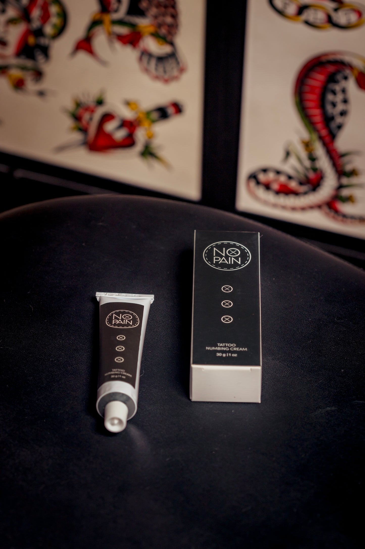 A tube and its corresponding box of "No Pain Tattoo Numbing Cream" from My Store are displayed on a dark surface. The box stands upright while the tube lies beside it, promoting pain-free tattooing. Both feature a minimalist black design with white text and icons, contrasted by colorful tattoo art in the background.