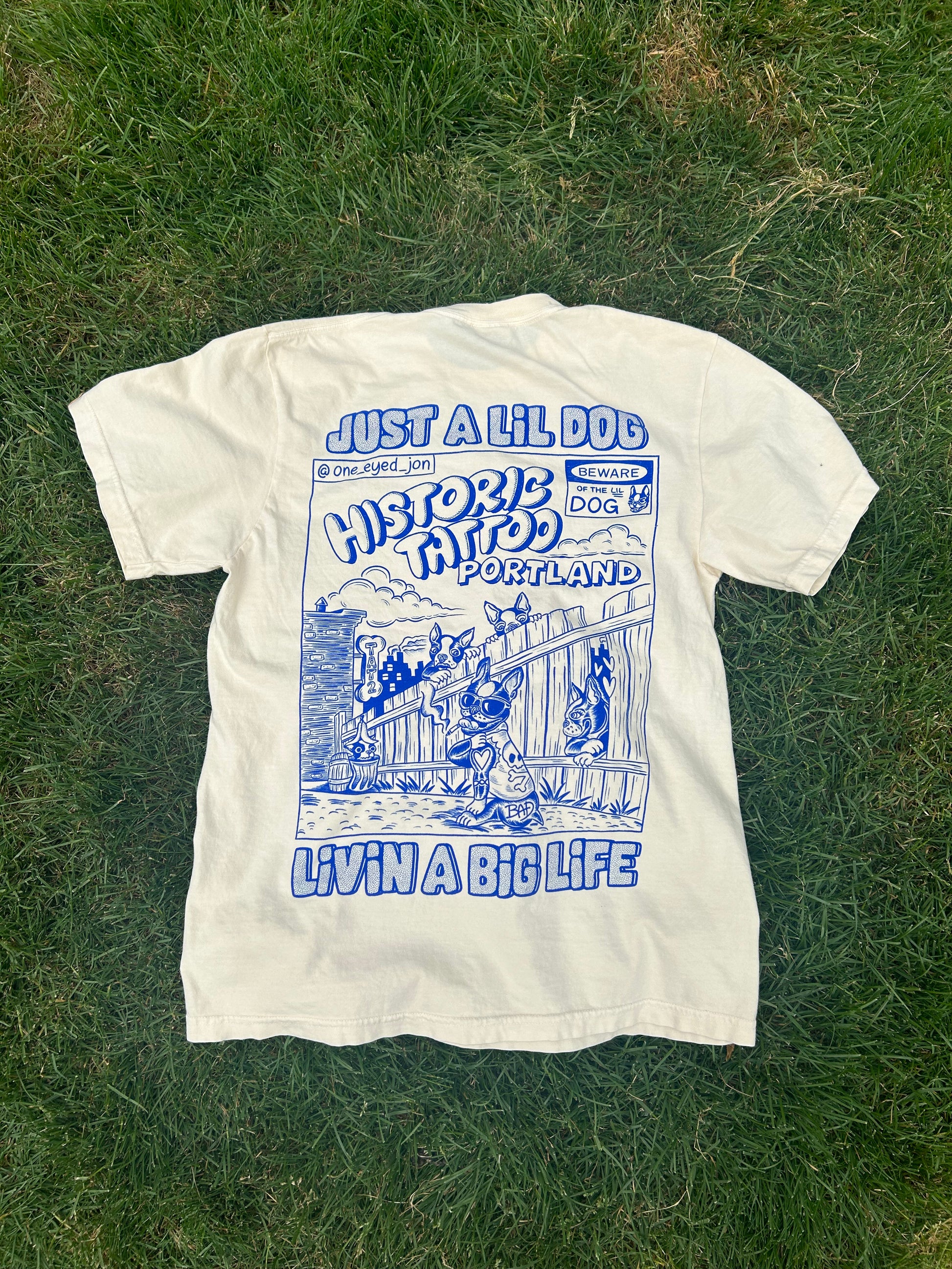 A Lil Dogs X Big Life T-Shirt on off-white Comfort Colors 100% cotton lies on grass with a blue graphic on the back. The design features an illustration of a dog in front of a tattoo shop, with text reading "Just a Lil Dog Historic Tattoos Portland Livin' a Big Life." This could easily be mistaken for a commemorative shirt from Boston. Sizes S-2XL T-Shirt by @one_eyed_jon.