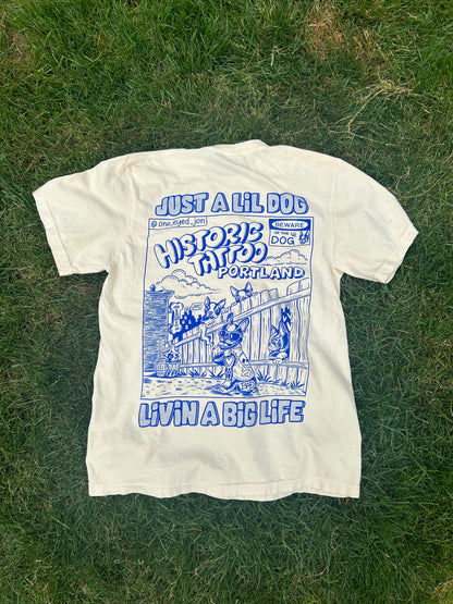 A Lil Dogs X Big Life T-Shirt on off-white Comfort Colors 100% cotton lies on grass with a blue graphic on the back. The design features an illustration of a dog in front of a tattoo shop, with text reading "Just a Lil Dog Historic Tattoos Portland Livin' a Big Life." This could easily be mistaken for a commemorative shirt from Boston. Sizes S-2XL T-Shirt by @one_eyed_jon.