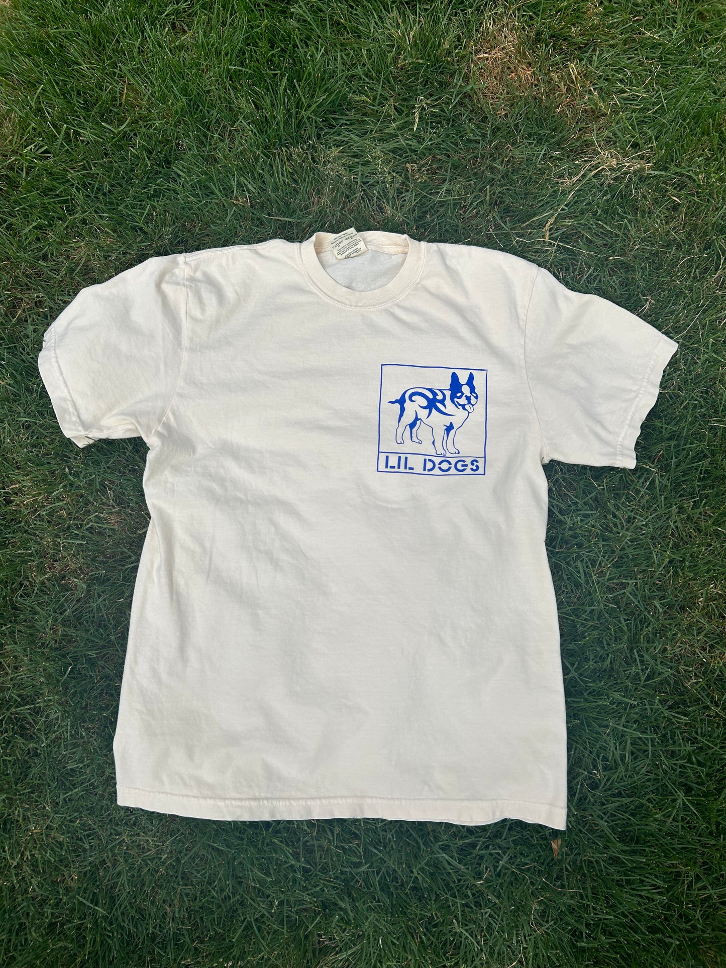 A Lil Dogs X Big Life T-Shirt on off-white Comfort Colors 100% cotton lies on grass, featuring a blue graphic of a dog inside a square on the left chest area. Below the graphic, text reads "LIL DOGS." This commemorative shirt captures the charm and playful spirit synonymous with Boston. Sizes S-2XL T-Shirt By @one_eyed_jon.