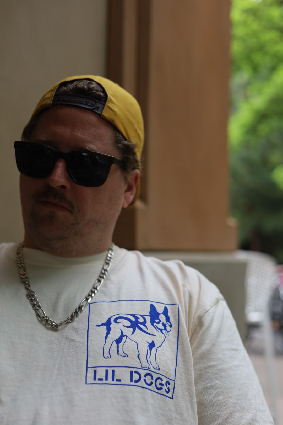 A man wearing a yellow baseball cap backwards, dark sunglasses, and a silver chain necklace. He has a short beard and mustache. Sporting historic tattoos on his arms, he also wears the Lil Dogs X Big Life T-Shirt on off-white Comfort Colors 100% cotton. Sizes S-2XL T-Shirt by @one_eyed_jon. The background shows an outdoor setting in Boston.