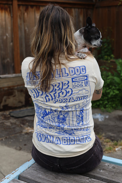 A person with long hair, wearing a Lil Dogs X Big Life T-Shirt on off-white Comfort Colors 100% cotton. Sizes S-2XL T-Shirt by @one_eyed_jon, is sitting outdoors. The shirt features the phrases “Just a Lil Dog” and “Livin' a Big Life.” The person holds a small black and white dog.