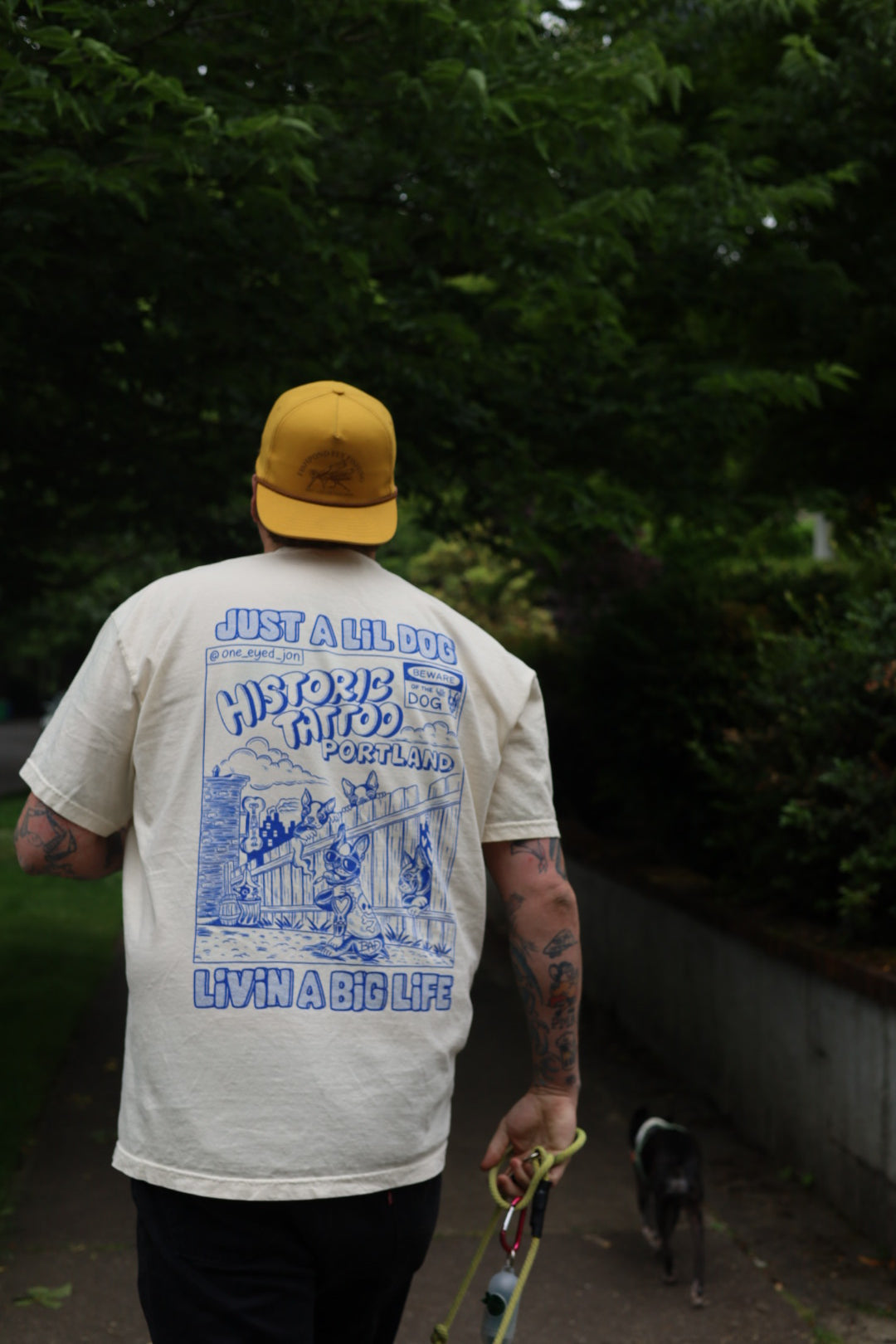 A person wearing a yellow hat and an off-white Lil Dogs X Big Life T-Shirt on off-white Comfort Colors 100% cotton, Sizes S-2XL T-Shirt By @one_eyed_jon with blue graphics and text on the back walks a small black and white dog. The commemorative shirt reads "Just a Lil Dog, Livin’ a Big Life." They stroll along a tree-lined path, surrounded by greenery and bushes in what looks like Boston.