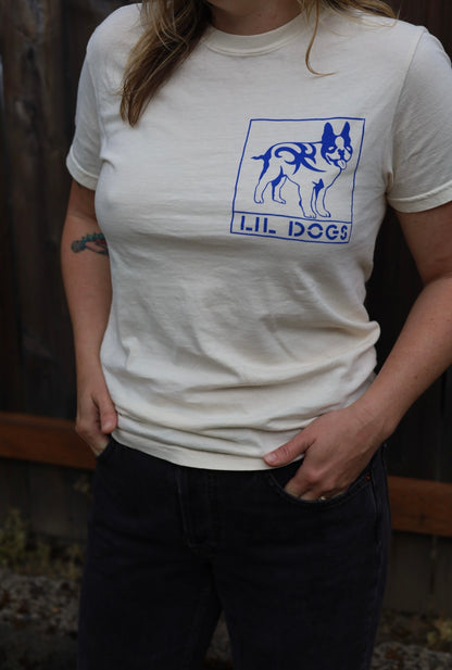 A person stands with hands in jean pockets, wearing a Lil Dogs X Big Life T-Shirt on off-white Comfort Colors 100% cotton. Sizes S-2XL T-Shirt By @one_eyed_jon. They have blonde hair and an intricate, historic tattoo on their left arm. A wooden fence is visible in the background.