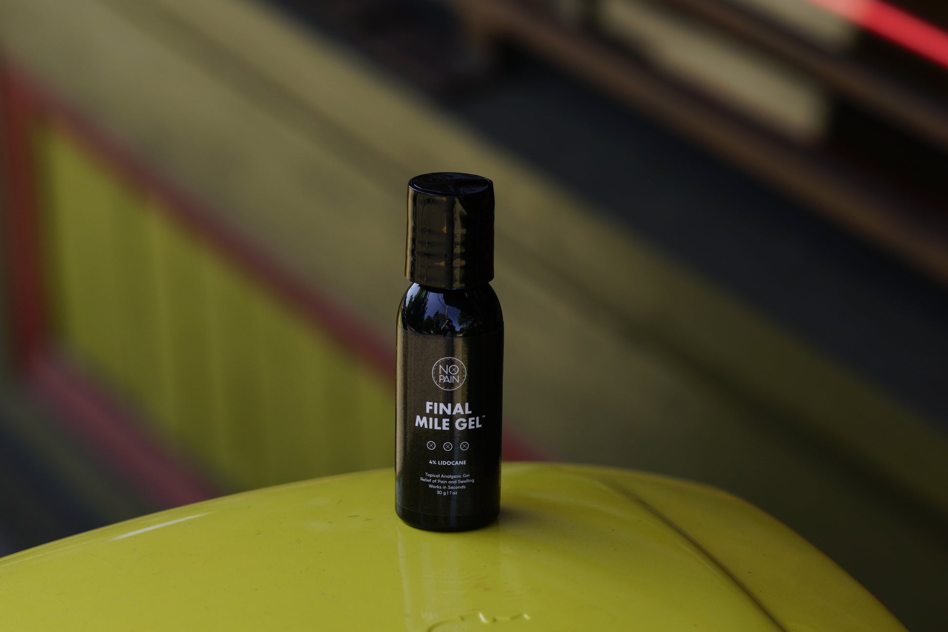 A small black bottle labeled "Final Mile Gel™ 4% Lidocaine" from No Pain Tattoo LLC sits on a smooth yellow surface, offering instant relief against a blurred background with some wooden elements and a hint of red trimming.