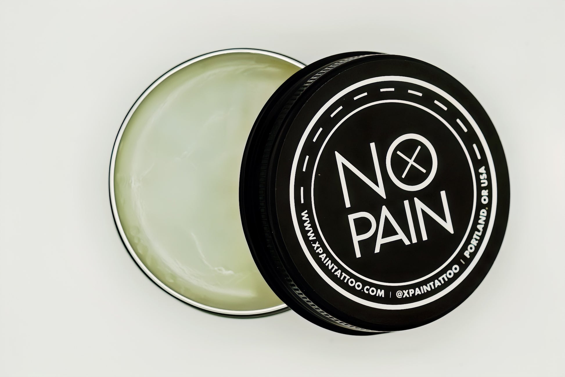 A black tin with a detachable lid, partly open, reveals the white No Pain Tattoo Aftercare Balm 30g inside. The lid displays the text "NO PAIN" along with a website URL and location information: "www.xpainattoo.com" and "Portland, OR USA." This soothing balm is an offering from My Store.