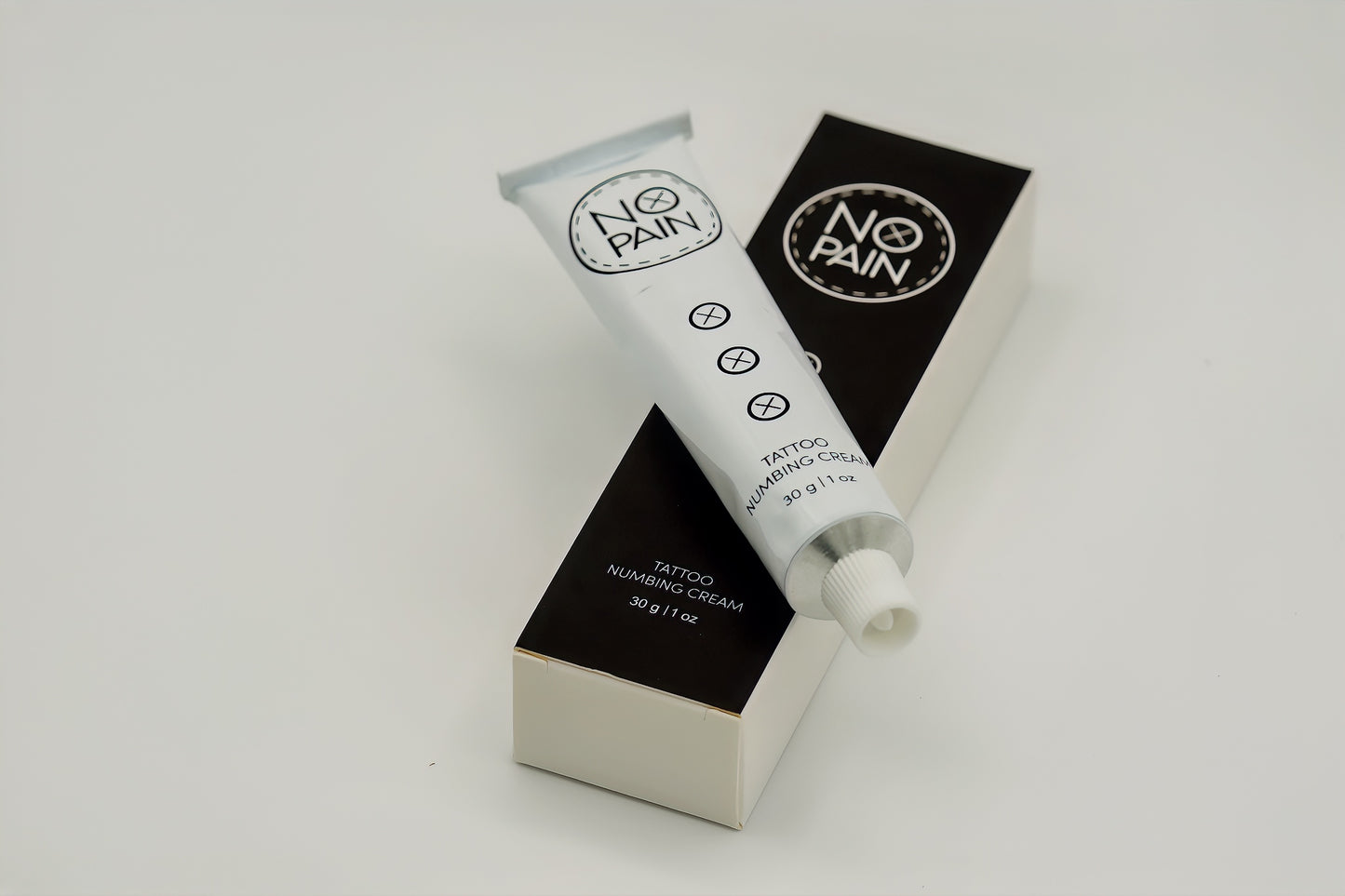 A white tube labeled "No Pain Tattoo Numbing Cream" from My Store rests on its black and white packaging box. The tube features a small open cap and displays product details, including "30g | 1 oz," along with symbols indicating it is designed for pain-free tattooing.