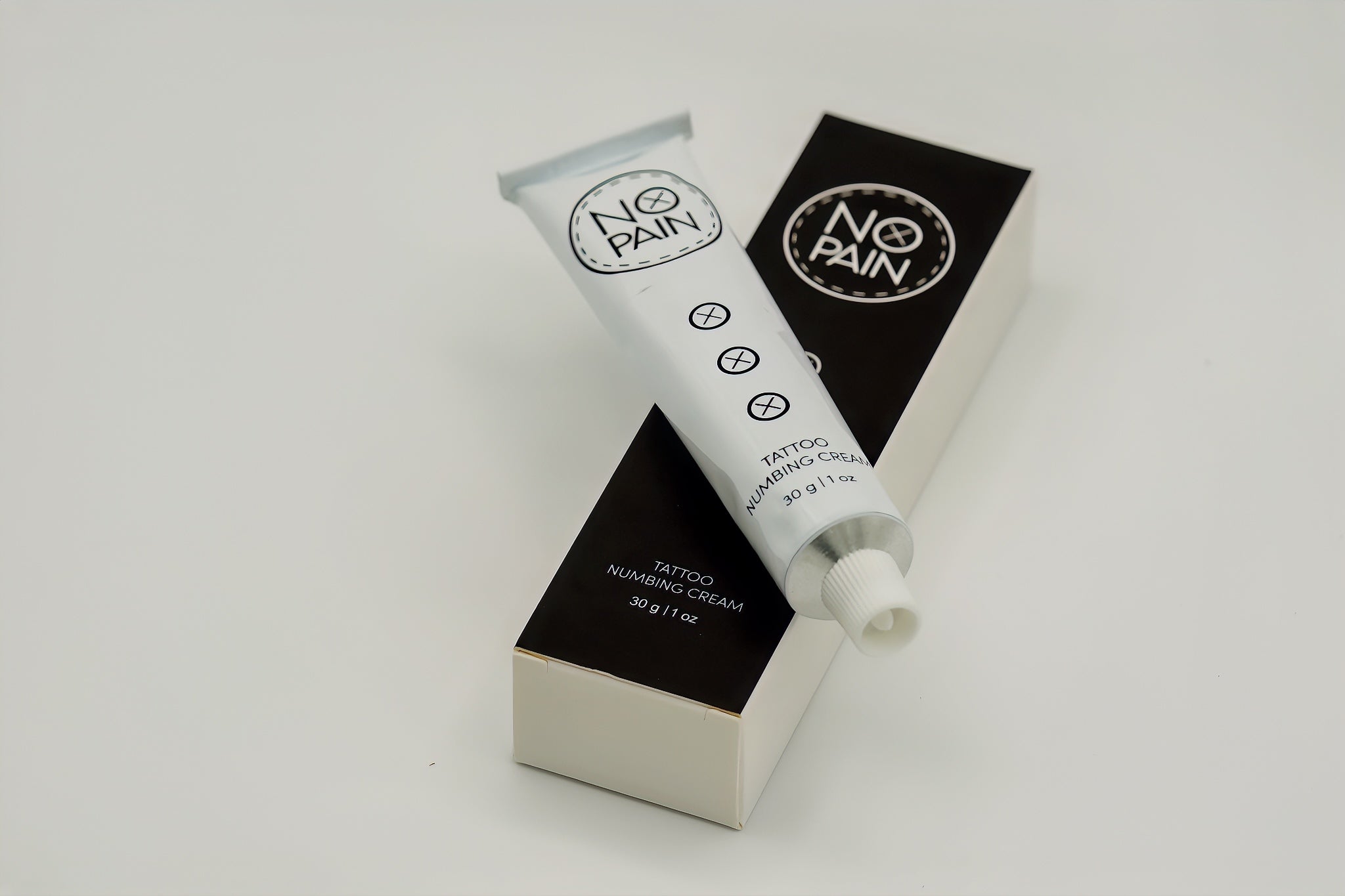 A white tube labeled "No Pain Tattoo Numbing Cream" rests on its black and white packaging box. The tube features a small open cap and displays product details, including "30g | 1 oz," along with symbols indicating it is designed for pain-free tattooing.
